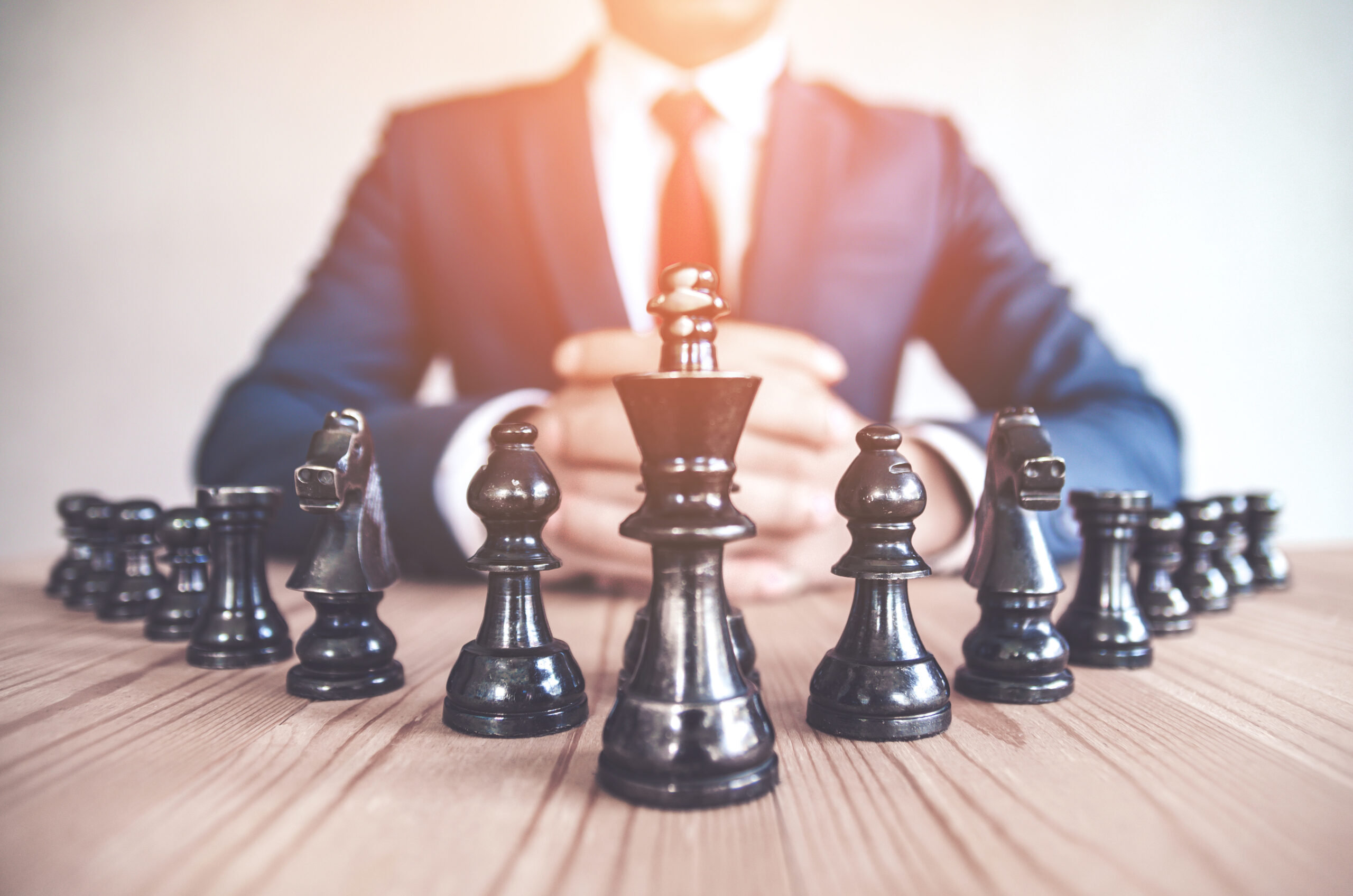 Retro style image of a businessman with clasped hands planning strategy with chess figures on an old wooden table. Prominence Global Marketing
